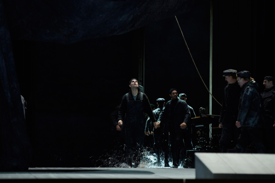 Der fliegende Holländer - © Thomas Jauk Der Steuermann tritt auf. Gekleidet in schwere Regenschutzkleidung, trägt er eine zarte Brille. Das Wasser, das auf der Bühne Lachen bildet, spritzt neben seinen Stiefeln hoch auf.