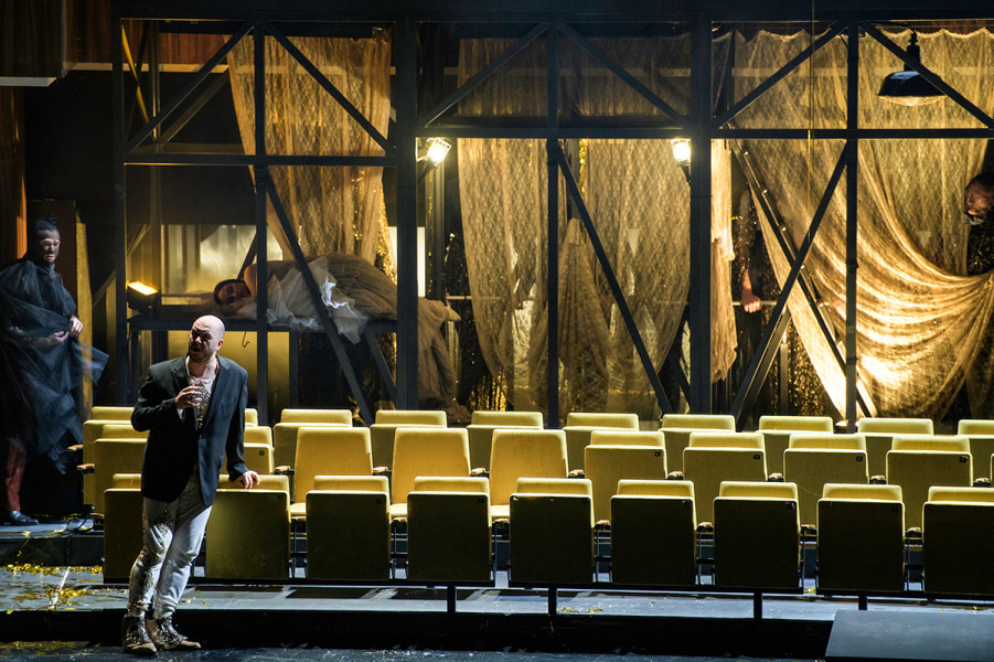 Rigoletto - © Bettina Stöß Während Rigoletto aus dem Unterbühnen-Mädchenzimmer vor die auf dem Boden verbliebenen vier Stuhlreihen tritt – er blickt dabei in Richtung Zuschauersaal, entführen hinter seinem Rücken maskierte Männer Gilda. Die Unterbühne fährt wieder ab.