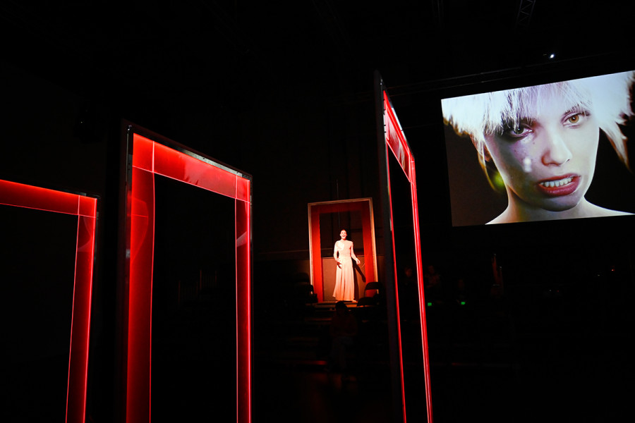 Das Bühnenbild von Beta besteht aus mehreren roten Türrahmen, die in verschiedenen Winkeln zueinander stehen können. Im oberen Türrahmen steht Scarlett, das Avatar des Medienmoguls Julian Zapp. Auf einem großen Bildschirm ist das Avatar verfremdet.