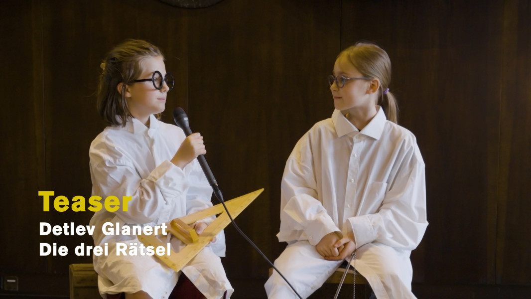 Die drei Rätsel – Ein Teaser - © Ruth Tromboukis Zwei Kinder aus dem Kinderchor sitzen im Foyer. Auf dem Knie des linken Mädchens ein großes Geodreieck. Beide Kinder tragen ihre "Alchimisten"-Brillen.