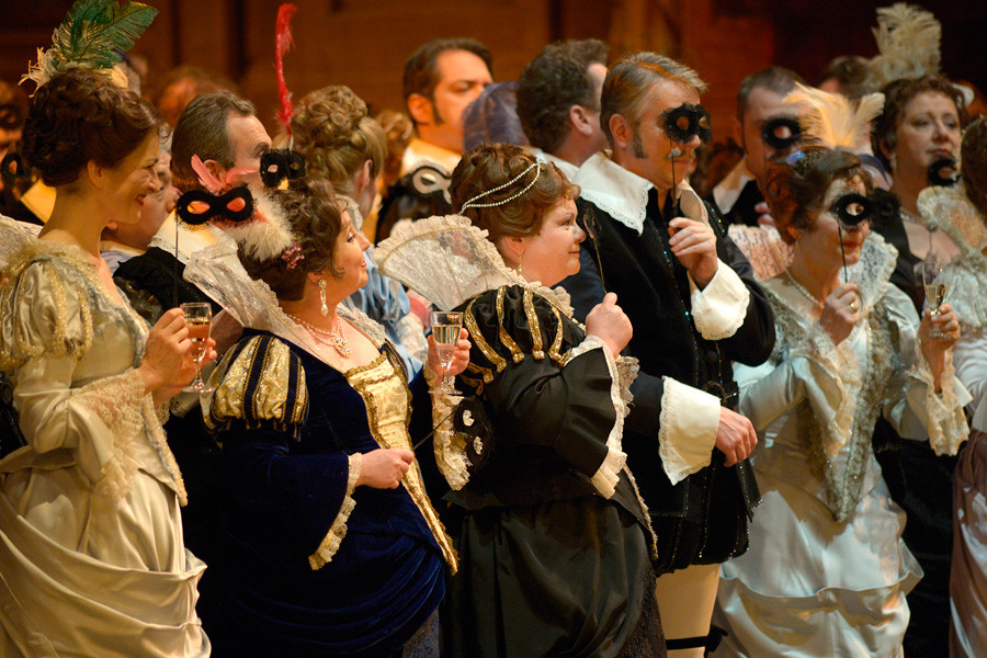 In historischen Kostümen (Samt und Seide) stehen die Damen des Chores. Vor die Augen halten sie Stabmasken.