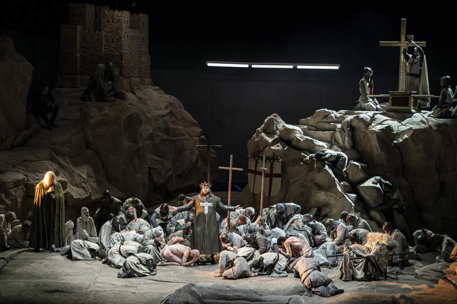 Parsifal - © Bettina Stöß Der Gral ist enthüllt: Auf dem Plateau rechts hebt der Priester das Gralsgefäß in die Höhe, Amfortas breitet seine Arme in Form eines Kreuzes aus, Titurel steht am Boden links in gleißendem Licht. Auf dem Burgberg haben sich Parsifal und Gurnemanz niedergelassen.