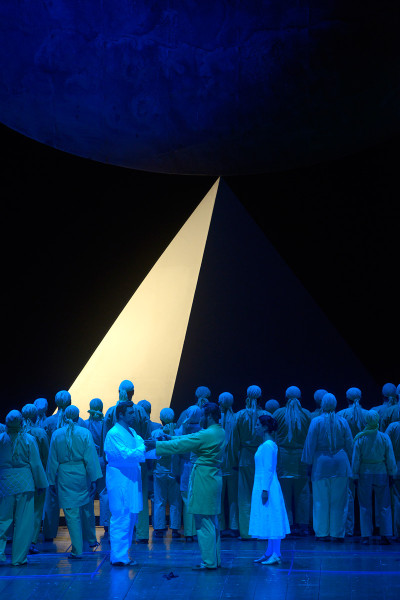 Die Zauberflöte - © Bettina Stöß Vor nachtblauem Horizont ragt eine golden angestrahlte Pyramide auf. Davor wandelt der Chor in seinen sonnengelben Gewändern. Sarastro begrüßt Tamino und Pamina.
