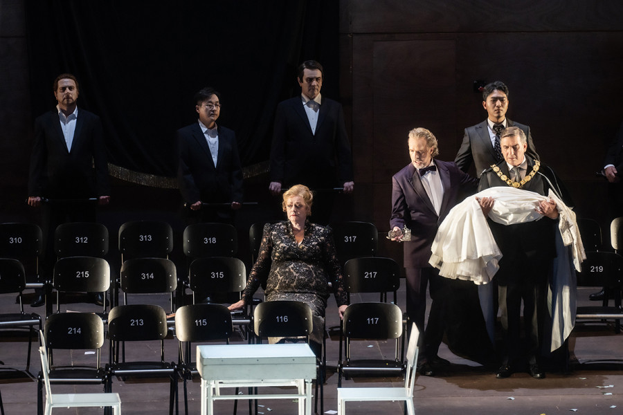 Turandot - © 2023 // Bettina Stöß Turandot sitzt in den leeren Stuhlreihen. Von hinten treten heran Calaf, der ihr Diadem in den Händen hält, und Altoum, Turandots Vater, mit einem Brautkleid über dem Arm.