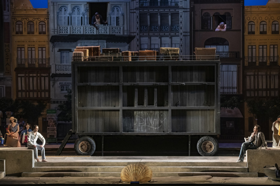 Il barbiere di Siviglia - © Bettina Stöß Auf diesem Foto ist eine Bühnentotale. Links und rechts sind kleine Mauervorsprünge, die die Straße vom Strand trennen. Jeweils dort sitzen Almaviva (links) und Figaro (rechts). Zwischen diesen Vorsprüngen hat der Theaterwagen geparkt. Dahinter sind die Fassanden einiger Häuser, die deutlich einen südeuropäischen Badeort imaginieren.