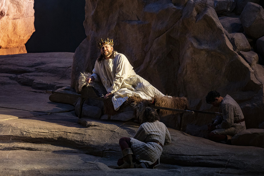 Parsifal - © Bettina Stöß König Amfortas wird von seinen Knappen zum Bade gebracht. Er sitzt auf einer Sänfte. Er ist gehüllt in einen Mantel, die Gralskrone auf dem Haupt. In sein Gesicht sind Leid und Schmerz eingegraben.