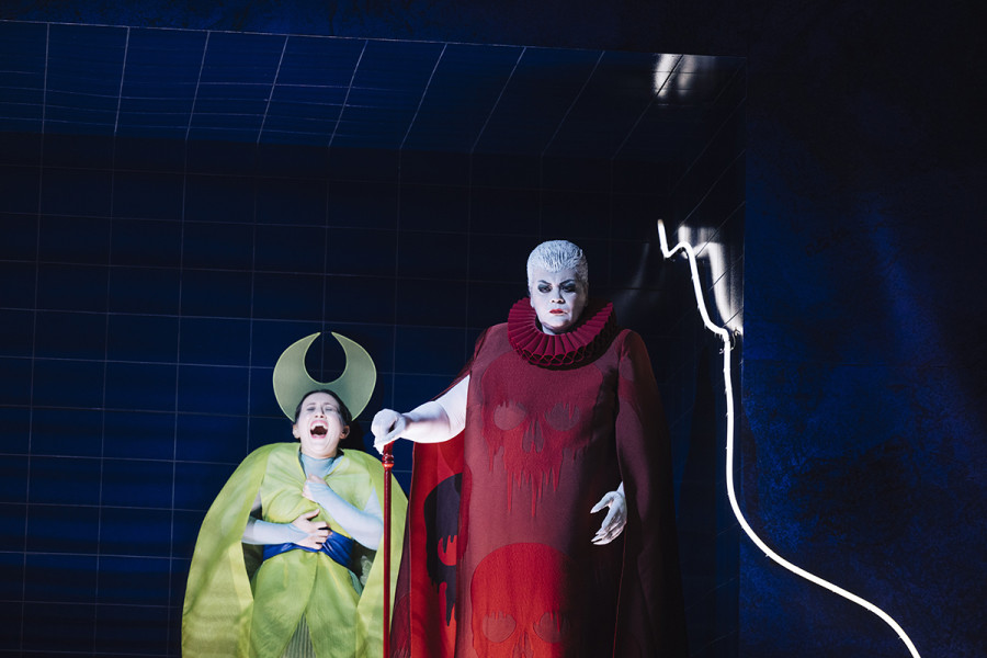 Suor Angelica - © Eike Walkenhorst La Zia Principessa schaut harten Blickes vor sich hin. Sie trägt ein langes rotes Kleid mit Totenköpfen. Die Haare sind sehr kurz, grau und nach hinten gebürstet. Sie hat einen roten Gehstock. Hinter ihr in schmerzverzehrendem Schreib Suor Angelica. Ihr Mantel ist goldgelb mit breiter blauer Schärpe unter der Brust. Auf dem Kopf trägt sie den halbmondförmigen Heiligenschein.