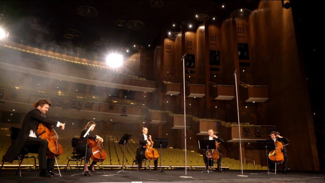 Fünf Cellist*innen sitzen auf der Bühne der Deutschen Oper Berlin, hinter ihnen der leere Saal der Deutschen Oper Berlin.