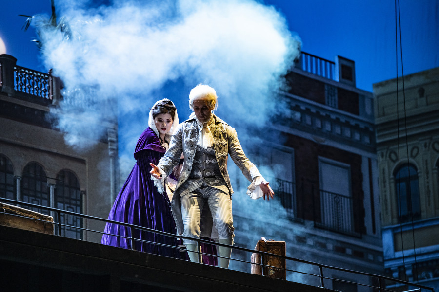 Dieses Foto ist eine Szenenimpression aus Katharina Thalbachs Inszenierung von IL BARBIERE DI SIVIGLIA (Der Barbier von Sevilla). Auf dem Dach eines Wohnwagens stehen Rosina und Almaviva. Im Hintergrund ist die Silhouette einer südlichen Stadt. Der Himmel ist nachtblau, vom Mond erhellt. Eine große Nebelwolke hat sich um die beiden Darsteller gebildet.