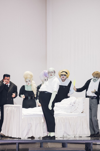 Die Darsteller*innen haben grün oder gelb geschminkte Gesichter und Hände. Die Kostüme sind schwarz/weiß. Die Verwandtschaft - Gherardo, Marco, Zita, Simone, Nella - umstehen das Bett des Buoso Donati. 