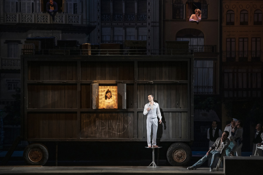 Il barbiere di Siviglia - © Bettina Stöß Rosina hat das Fenster ihres Wohnwagens geöffnet. Auf einem kleinen Kaffeehaus-Tischchen steht Almaviva, sein Ständchen singend. Figaro hat sich über sein Hemd noch eine Lederjacke gezogen und sitzt etwas rechts der Szenerie.