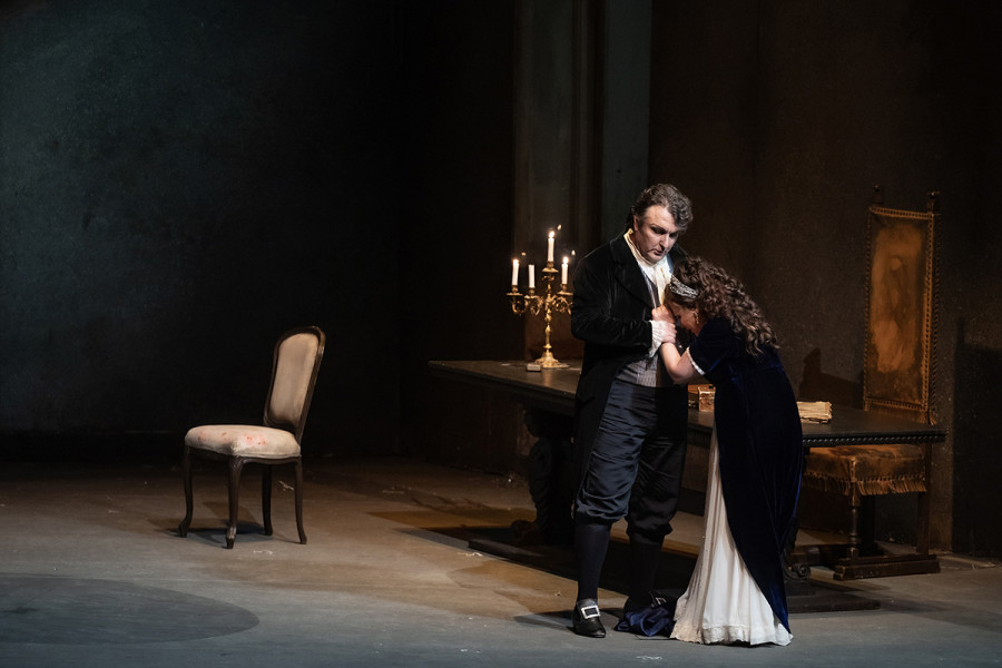 Tosca - © 2023, Bettina Stöß Tosca und Scarpia stehen am Schreibtisch. Scarpia hält Toscas Handgelenke fest umschlossen. Tosca lässt flehend den Kopf gegen Scarpias Brust sinken.