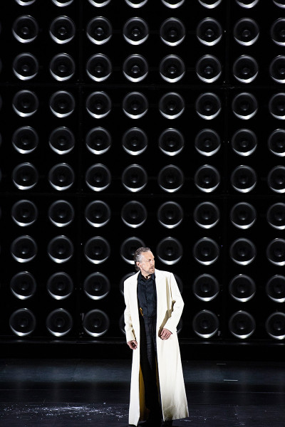 Tristan und Isolde -   © Bernd Uhlig  Die Lampen sind vollständig erloschen. Vor der Wand steht König Marke in schwarzem Hemd und schwarzer Hose und einem weißen bodenlangen Mantel.