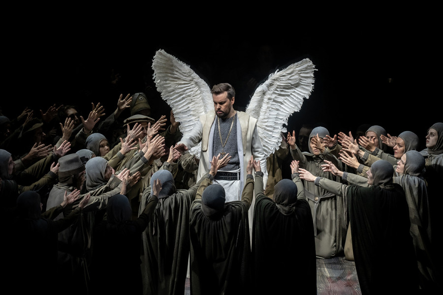 Lohengrin - © Bettina Stöß Lohengrin steht mit seinen Engelsflügeln mitten im Chor. Die Männer und Frauen knien teilweise und heben die Hände flehend zu ihm auf.