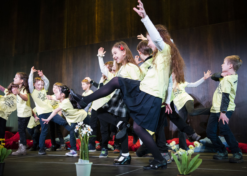 Frühlingssingen - © S.I. Der Kleine Chor des Kinderchores bei seinem Frühlingssingen: Die Kinder singen und tanzen auf der Foyerbühne in hellgelben T-Shirts. Verstreut sind einige Narzissen und Frühblüher.