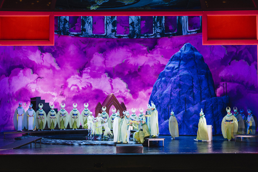 Suor Angelica - © Eike Walkenhorst Bühnentotale mit allen Bestandteilen: Ein blauer Felsen, die orangene Kapelle, eine Treppe und das Wasserbecken. Verschiedene Gruppen von Nonnen verteilen sich in diesem Bild.
