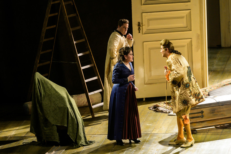 Le nozze di Figaro - © Bettina Stöß Don Basilio stört das "Zusammensein" von Almaviva und Susanna. Almaviva beherrscht nur mühsam seine Wut. Basilio macht die Entdeckung der beiden sichtlich Spaß. Im Lehnstuhl unter einer grünen Decke kann man Cherubino vermuten. Don Basilio trägt stark onduliertes Haar, weißseidene Kniebundhosen, orange Strümpfe, Schnallenschuhe und einen bestickten Rock.