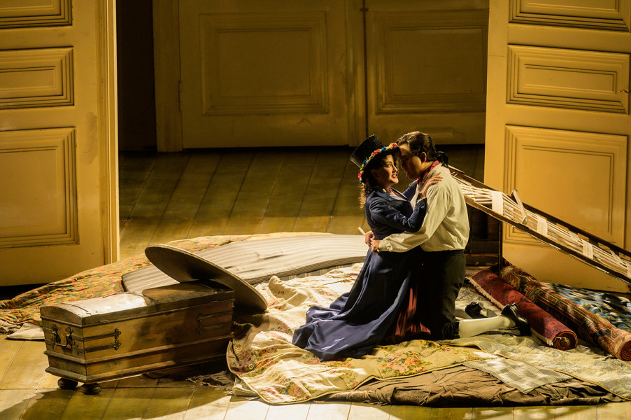 Le nozze di Figaro - © Bettina Stöß Figaro und Susanna knien auf ihrer zukünftigen Ausstattung. Teppiche, ein Bettkasten, eine Truhe, ein Spiegel ... Er trägt ein weißes Hemd und ein rotes Halstuch, sie ein blaues Kleid und einen blumenbekränzten Zylinder. Eine Flügeltür hinter ihnen ist weit geöffnet.