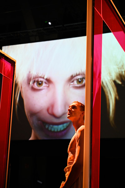Der Medienmogul Julian Zapp lehnt an einem rot beleuchteten Türrahmen, hinter ihm strahlt sein Avatar Scarlett auf einem Bildschirm.