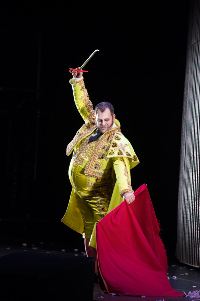 Carmen - © Marcus Lieberenz Der Matador Escamillo schwingt in einem sehr auffälligen, gold-gelben Torero-Kostüm seine rote "capote" (das zum Stierkampf gehörende Tuch). In seiner rechten Hand hält er ein Messer.