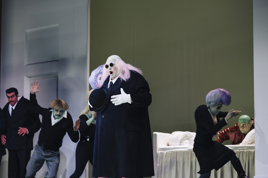Gianni Schicchi - © Eike Walkenhorst Der Arzt, der Buoso besuchen kommt, und von der Verwandtschaft und Gianni Schicchi betrogen wird, in einem schwarzen Mantel, langem grauen Haar, grünlichem Gesicht und Sonnenbrille. Um ihn herum sind alle in heller Aufregung. Gianni kniet listig hinter dem Bett.