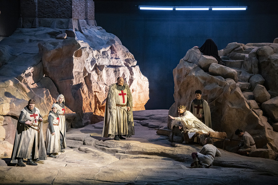 Parsifal - © Bettina Stöß Umgeben von den Knappen und Gralsrittern Gurnemanz und Amfortas. Gurnemanz, die Knappen und die Ritter tragen Kostüme, die an die mittelalterlichen Kreuzritter angelehnt sind. Oben auf dem Plateau sitzt in einen schwarzen Umhang gehüllt Kundry.