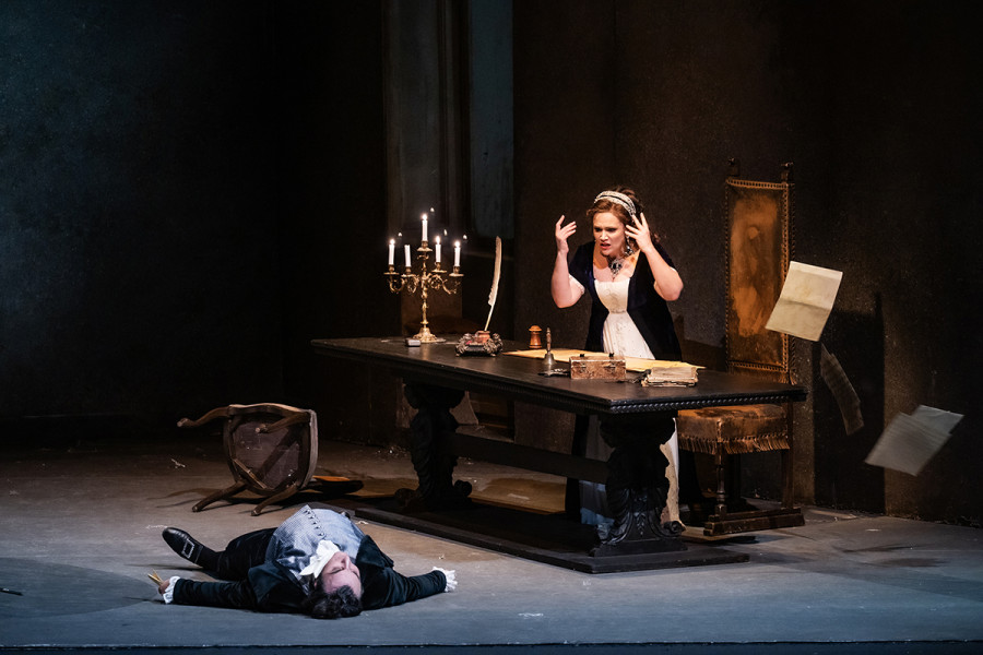 Tosca - © 2023, Bettina Stöß Scarpia liegt - von Tosca erstochen - tot am Boden. Tosca ist hinter Scarpias Schreibtisch, ein Dokument suchend, das ihr und Cavaradossi freien Weg aus Rom gewähren soll. Sie gewahrt, dass Scarpia dies in seiner linken Hand hält.