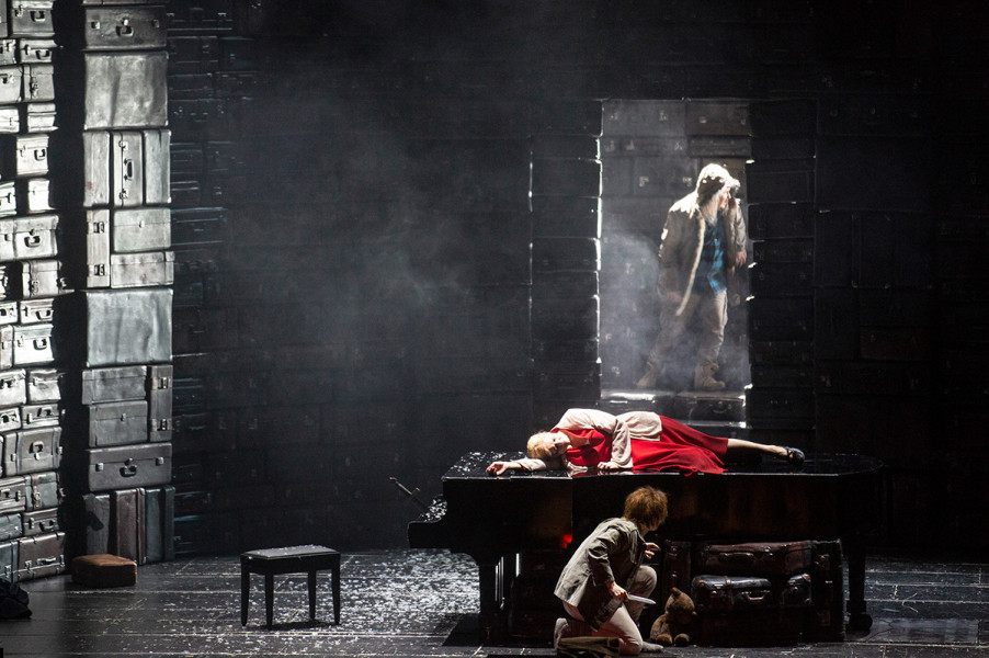 Die Walküre - © Bernd Uhlig Auf der Flucht erreicht Siegmund verwundet Hundings Haus. Siegmund steht im Nebel in der Tür. Auf dem Flügel liegt ruhend im roten Kleid Sieglinde. Hundingling, der gemeinsame und ungeliebte Sohn von Sieglinde und Hunding, verbirgt sich vor dem Fremden unter dem Flügel.