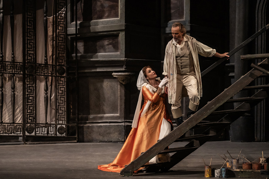 Tosca - © 2023, Bettina Stöß Tosca kniet auf der unteren Stufe einer Treppe, die zu dem Fresko führt, an dem Cavaradossi gerade arbeitet. Flehend blickt sie auf Cavaradossi, dessen Hand sie hält.