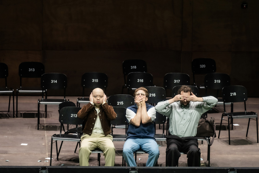 Turandot - © 2023 // Bettina Stöß Zwischen den leeren Stuhlreihen sitzen Pong, der sich die Ohren zuhält, Pang, der sich den Mund zuhält und Ping, der sich die Augen bedeckt.