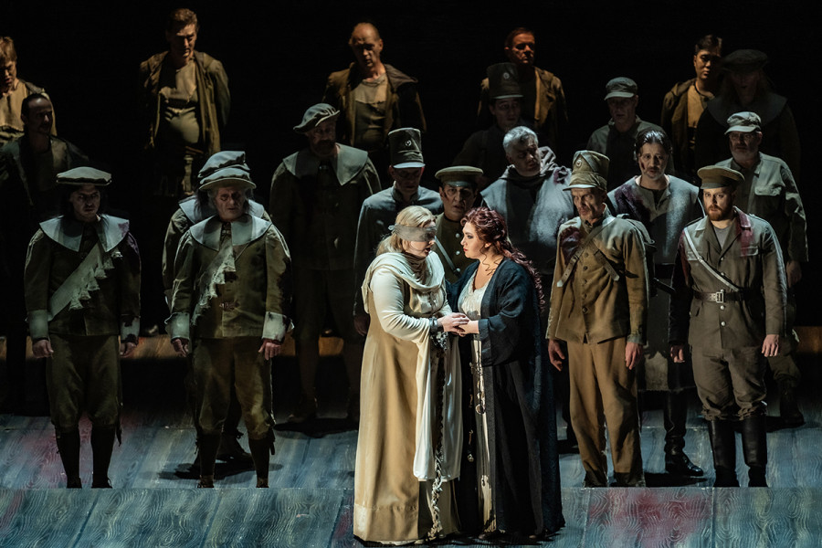 Lohengrin - © Bettina Stöß Ortrud stützt Elsa, deren Augen durch ein Tuch verhüllt sind. Hinter ihnen stehen die Herren des Chores. Die Kostüme des Herrenchores sind blutbefleckt, die Gesichter sind leichenhaft blass und oft mit schweren Wunden übersät.