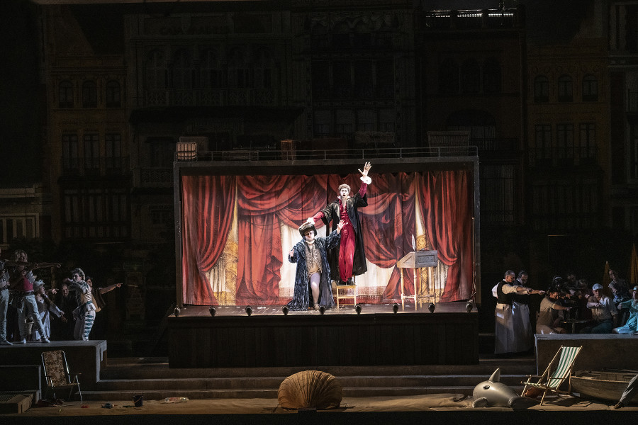 Il barbiere di Siviglia - © Bettina Stöß Wieder eine Bühnentotale: Links und rechts hinter den Mauervorsprüngen ist nun quicklebendiges Abendleben, im Wohnwagen selbst vor dem gemalten Spielvorhang Bartolo mit Don Basilio. Basilio hat ein feuerrotes Gewand mit einem schwarzen Mantel, einem übergroßen Hut und "verlängerten Händen", will heißen: Er trägt über seinen Händen Handschuhe, die die Finger stark verlängern.