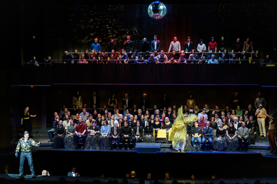 Der Herzog steht auf der Passerelle. Auf der Bühne ist der Saal  der Deutschen Oper Berlin spiegelverkehrt aufgebaut. Der Chor und die Statisterie sitzen in den  Stuhlreihen, genauso wie Rigoletto in seinem goldenen Lametta-Hasen-Kostüm.
