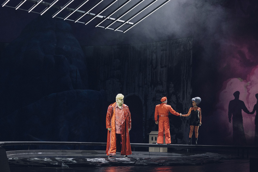Eine Szene aus IL TABARRO: Michele in rotem Mantel und rotem Kostüm steht mit bösem Blick in der Mitte der Bühne. Am rechen Rand das Pärchen der Due Amanti.