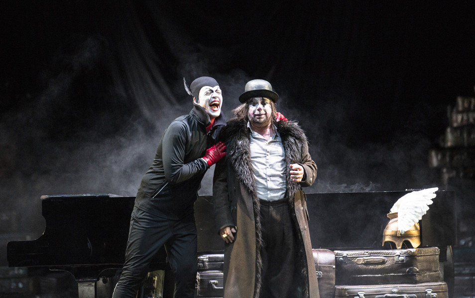 Das Rheingold - © Bernd Uhlig Loge lacht schallend neben Alberich. Loge trägt ein Kostüm, das an ein klassisches Mephisto-Kostüm erinnert, Alberich trägt Melone, weißes Hemd, eine schmutzige schwarze Hose und einen abgerissenen Leder-Fell-Mantel.
