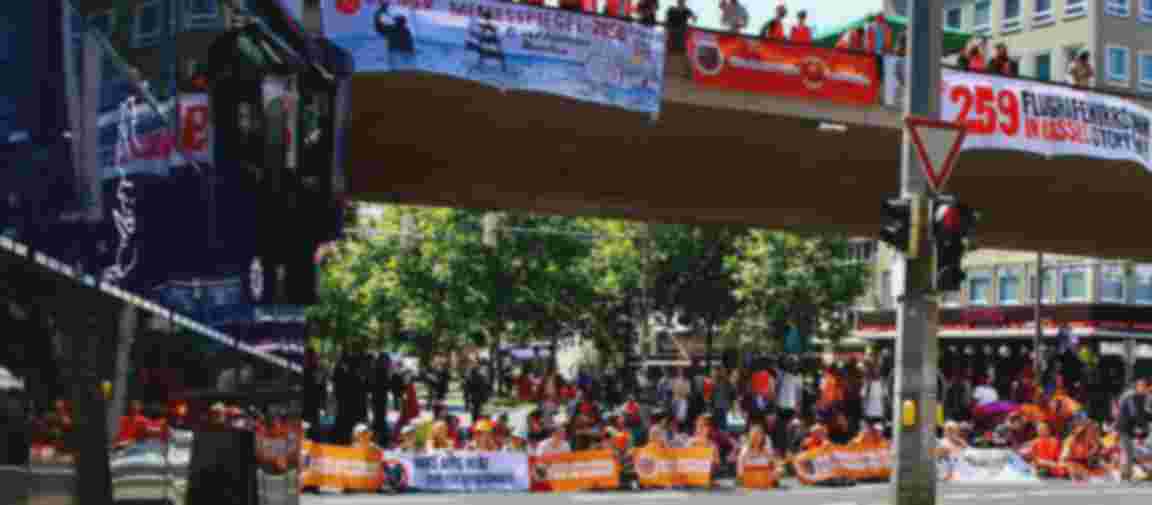 Auf einer Straße sitzen viele Menschen mit orangenen Bannern. Auch von der Brücke über den Menschen hängen Banner mit politischen Botschaften. Auf einer Straße sitzen viele Menschen mit orangenen Bannern. Auch von der Brücke über den Menschen hängen Banner mit politischen Botschaften.
