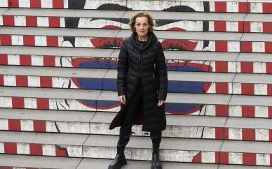 Christiane Mudra steht, gekleidet in schwarzen Parka und schwarze Stiefel, auf einer Treppe, auf deren Stufen ein Graffiti ist mit weit geöffneten Augen in rot und blau.