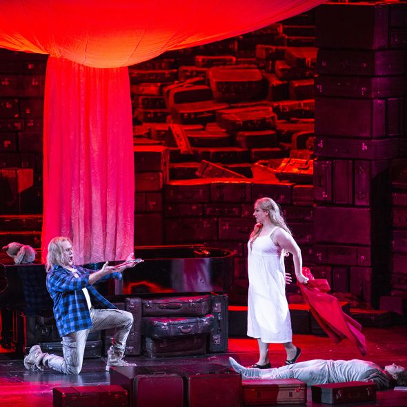 Eine Szenenimpression aus DIE WALKÜRE: Siegmund kniet vor Sieglinde, die auf ihn zugeht. Den Hintergrund bilden Koffer. Die Szenerie ist in rotes Licht getaucht.
