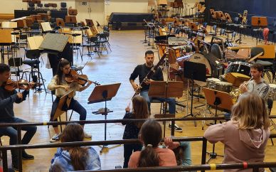 Der Blick geht von der Chorempore in den Orchesterprobensaal mit Musikern der Deutschen Oper Berlin, in der Mitte ein Fagottist. Von hinten sieht man die Kinder dem Instrument lauschen.