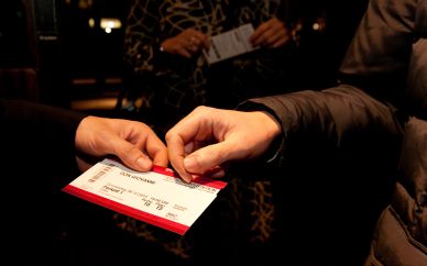 Im Mittelpunkt des Bildes ist eine Eintrittskarte und zwei Hände. Fotografiert wurde eine typische Einlass-Situation, bei der der Besucher das Ticket präsentiert und der Kontrolleur es entwertet.