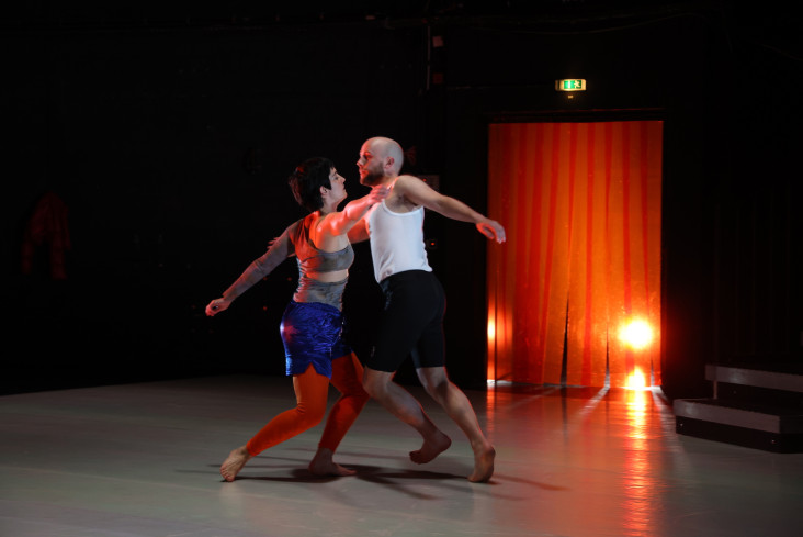 Vor einem Vorhang aus großen Plastiklamellen tanzen zwei Personen miteinander. Der Vorhang wird von hinten angestrahlt und leuchtet deswegen orange.