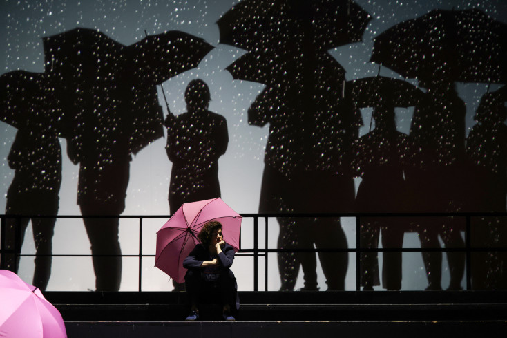 Eine Sängerin sitzt auf einer Stufe unter einem rosa Regenschirm. Hinter ihr sind große Schatten von Menschen mit Regenschirmen zu erkennen.