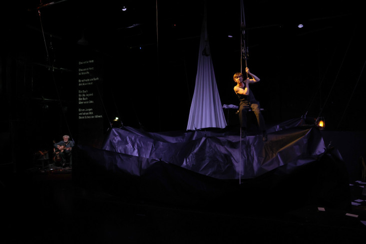 Die Bühne ist dunkel und voller schwarzem Papier. Hinten spielt ein Mann Gitarre, ein anderer Mann sitzt auf einer Art Schaukel in der Luft.
