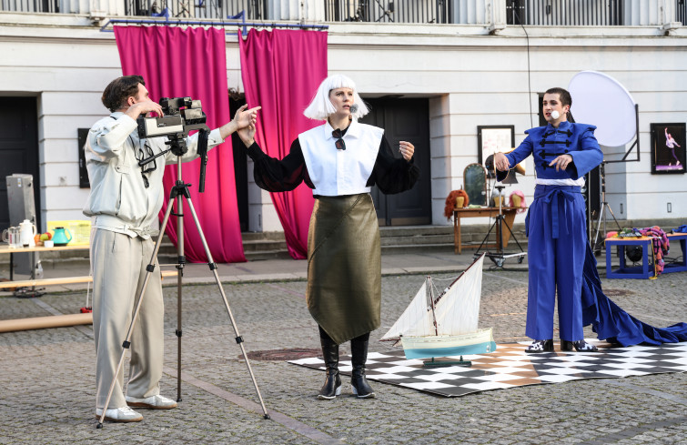 Auf dem Goetheplatz stehen drei Personen. Ein Schauspieler ist ein Kameramann. Er filmt eine Schauspielerin. Sie trägt eine weiße Perücke und schwarz-weiße Kleidung. Hinter ihr steht ein Schauspieler in einem blauen Kostüm. An der Front des Theaters hängt ein großer, roter Vorhang.