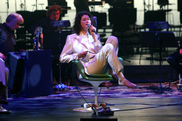 Eine Sängerin sitzt mit angezogenen Beinen im Drehstuhl und singt. Ihre Schuhe stehen auf dem Boden. Die Band spielt hinter ihr.