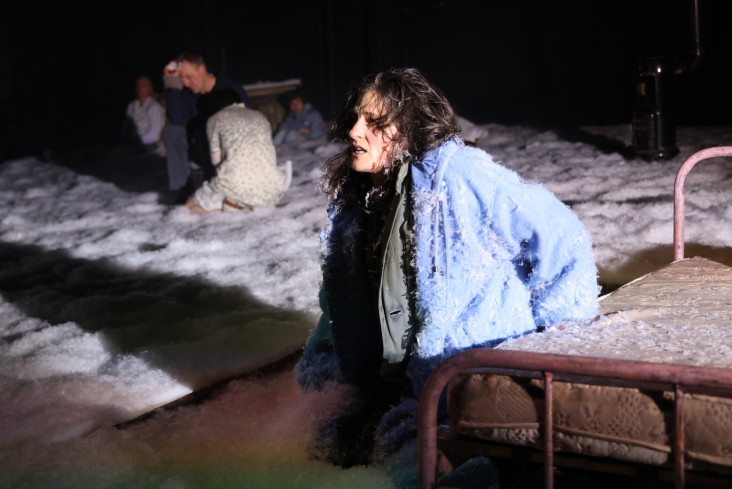 Neben einem Bett kniet eine Schauspielerin. Sie trägt eine Winterjacke. Der Bühnenboden ist mit Kunstschnee bedeckt.