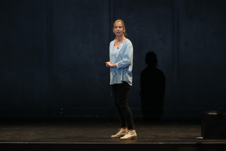 In schwarzer Hose, blauem Oberteil und weißen Schuhen steht die Schauspielerin seitlich auf der Bühne und spricht ins Publikum.