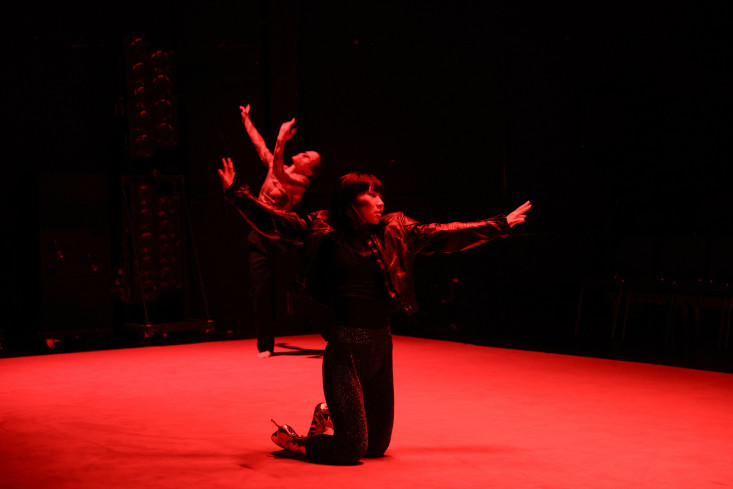 Eine Tänzerin kniet im Schweinwerferlicht auf dem roten Teppich. Sie hat die Arme ausgebreitet und ist ganz schwarz angezogen.