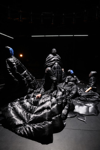 Auf der schwarzen Bühne stehen und sitzen fünf Personen zusammen. Sie tragen alle lange, schwarze Kleider, ihre Gesichter sind verhüllt. 