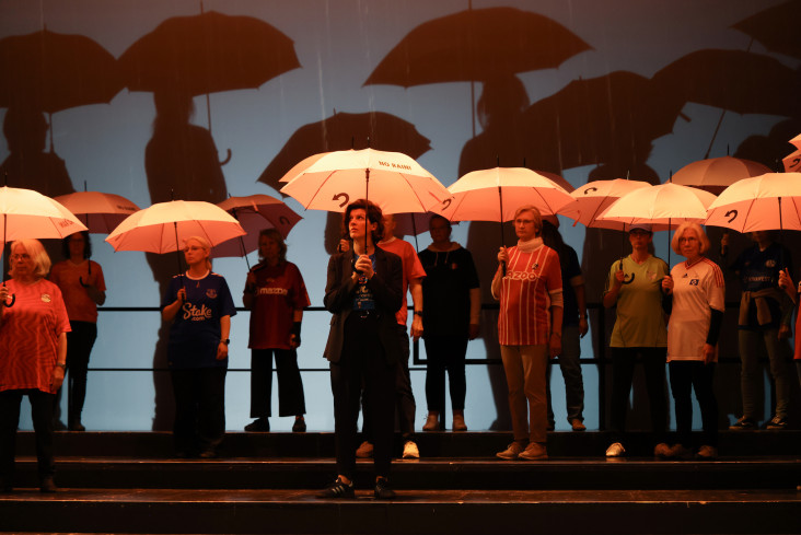 Viele Menschen mit aufgespannten Regenschirmen stehen auf der Bühne. 